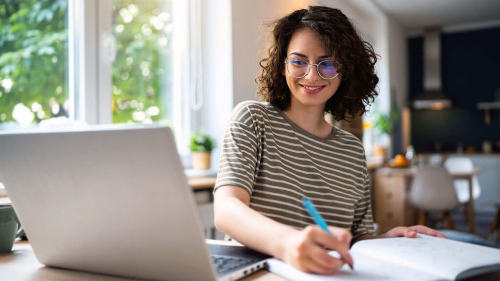 a woman wearing glasses gazes at her laptop while taking notes in a notebook